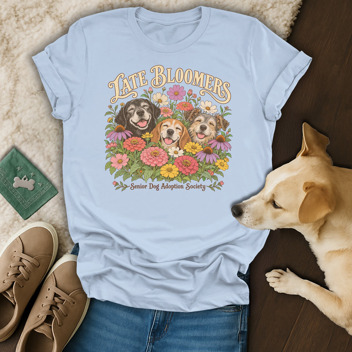 A light blue t-shirt with a graphic of three dogs surrounded by flowers and the text Late Bloomers Senior Dog Adoption Society, laid out flat with a live dog, shoes, and a wallet on the side.