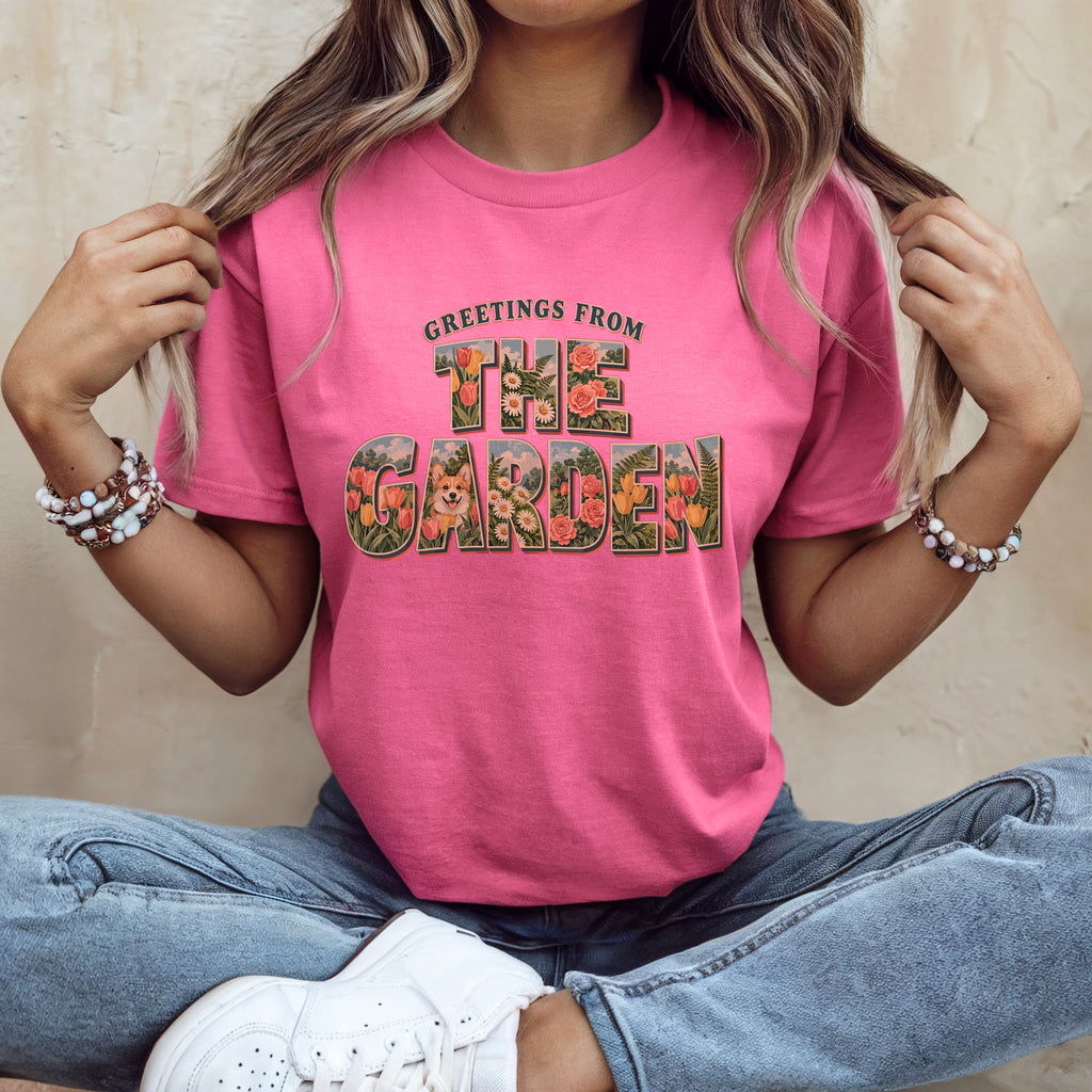 A person wearing a pink t-shirt with Greetings From The Garden graphic text with a corgi and floral details.