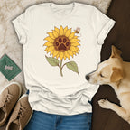 A cream-colored t-shirt featuring a sunflower with a paw print center and a bee, lying on jeans next to a dog, shoes, and a green passport holder.