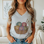 A woman wearing a gray V-neck t-shirt featuring a graphic of yarn balls with paw prints in a basket.
