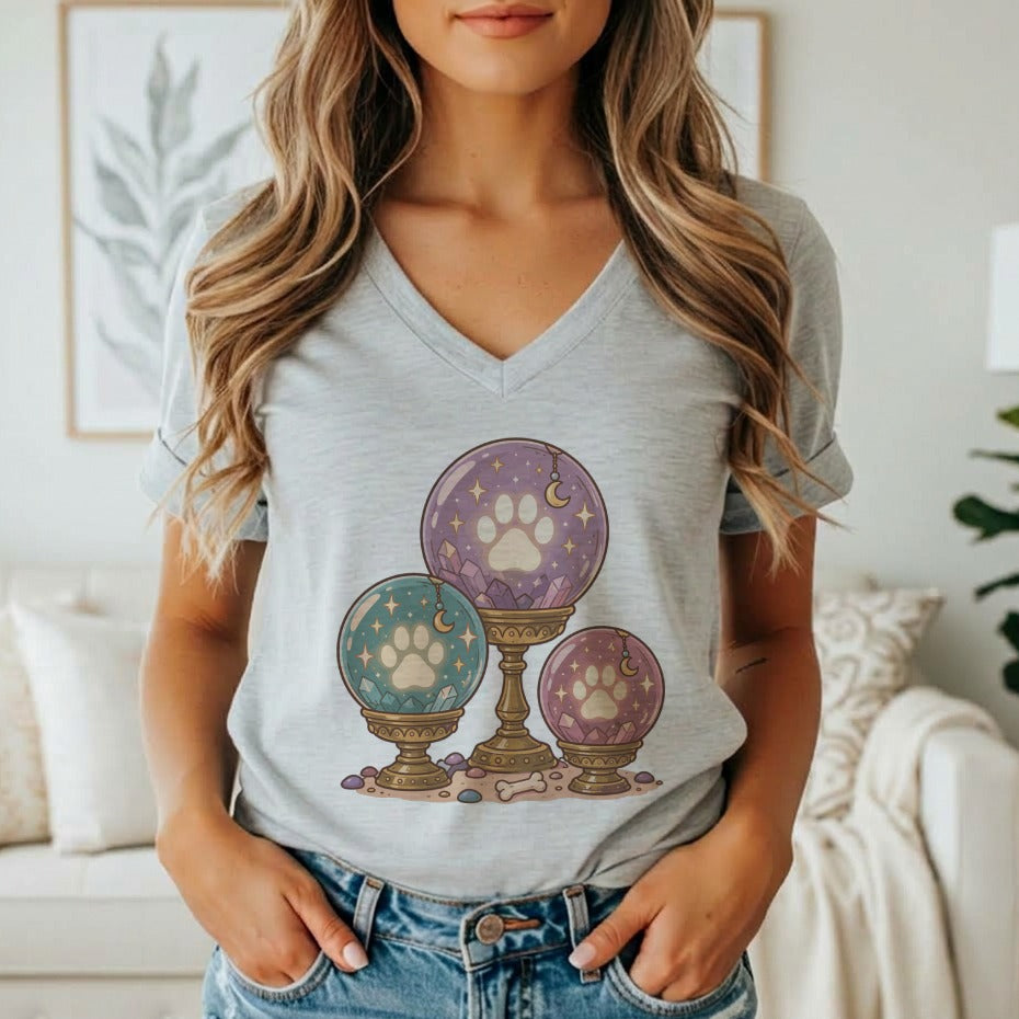 A woman wears a grey V-neck t-shirt featuring a graphic of three glowing paw print crystal balls.