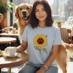 A smiling woman with dark hair wearing a light blue t-shirt with a sunflower graphic while a golden retriever stands beside her.