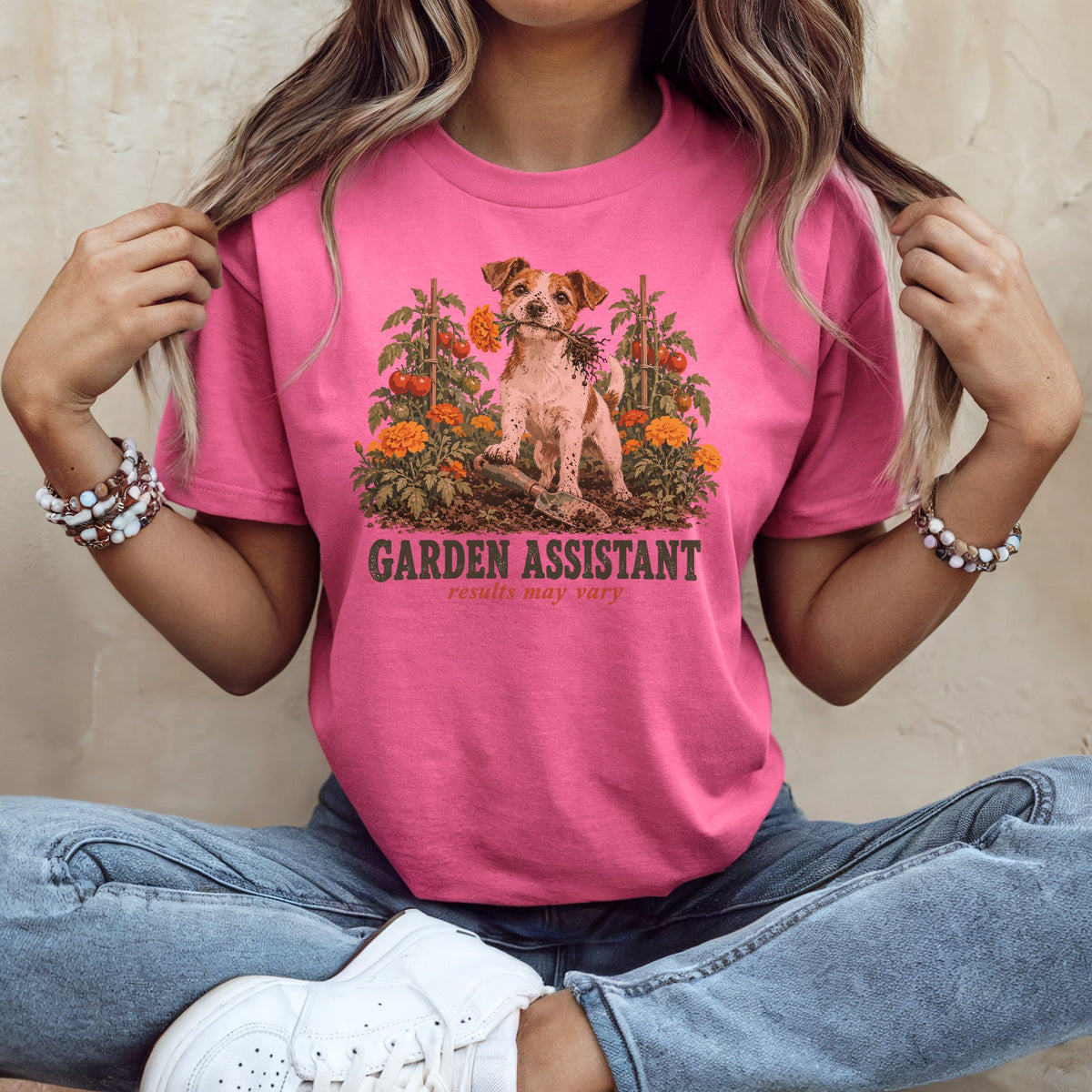 A person sitting cross-legged wearing a pink crewneck t-shirt featuring a dog surrounded by garden plants and the text Garden Assistant.