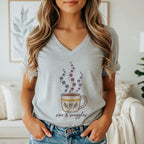 A woman wears a grey v-neck t-shirt featuring a coffee mug graphic with paw print steam and the text sips and snuggles.
