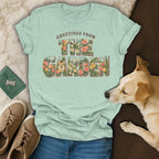 Light green t-shirt featuring floral text saying Greetings From The Garden, displayed with jeans, sneakers, and a dog.