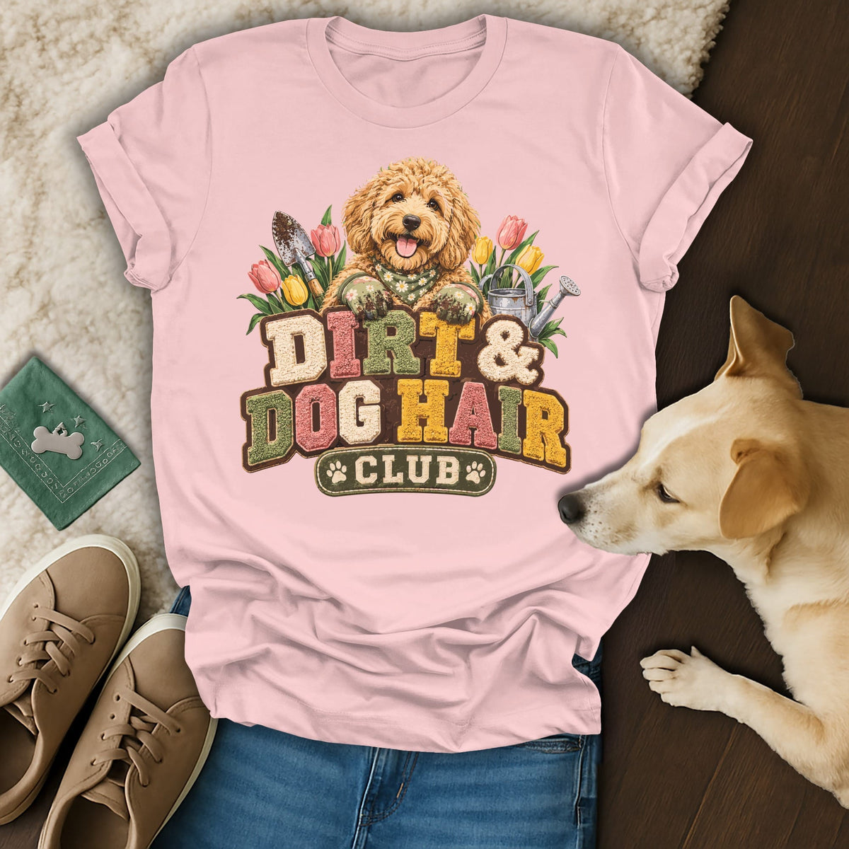 A light pink t-shirt with a graphic of a golden doodle, gardening tools, flowers, and the text "Dirt and Dog Hair Club."