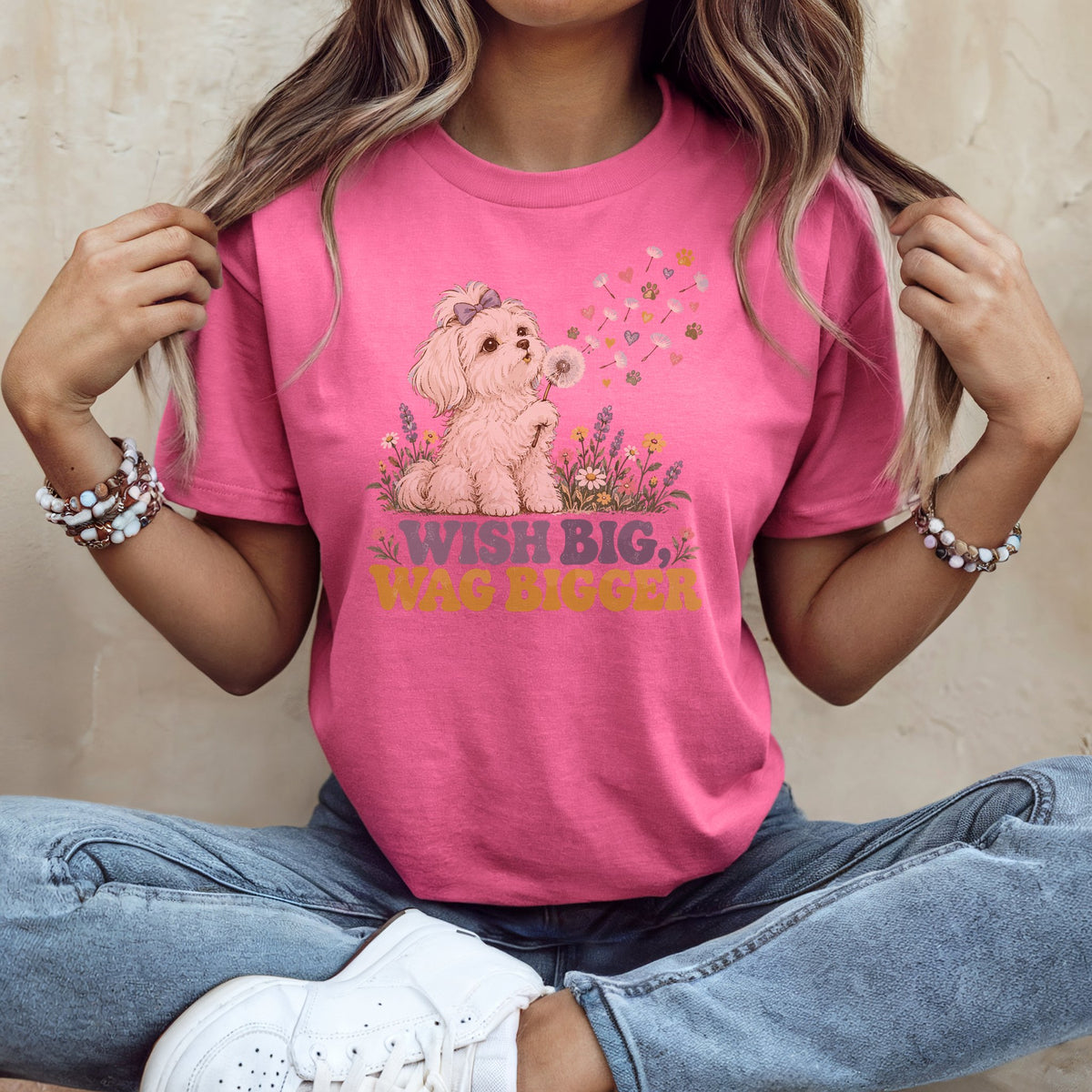 A woman wearing a pink t-shirt with a graphic of a puppy blowing dandelion seeds and the text Wish Big, Wag Bigger.