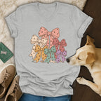 Grey V-neck t-shirt featuring a graphic of colorful bows printed with paw prints, laid out with a dog and sneakers.