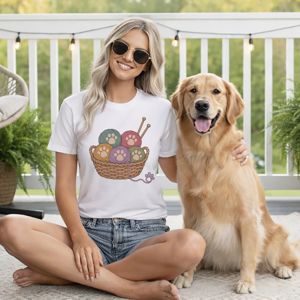 A smiling blonde woman wearing sunglasses and a yarn-basket t-shirt sits with a golden retriever outdoors.