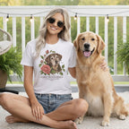A smiling woman wearing a white t-shirt featuring a dog and rose graphic sits next to a golden retriever.