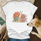 A white V-neck t-shirt featuring a cartoon illustration of colorful snails with paw print shells, laid flat next to sneakers and a dog.
