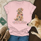 A light pink t-shirt featuring a patchwork dog graphic and the text "PERFECTLY PATCHED" lies flat on a textured surface with accessories and a real dog.