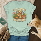 A light green t-shirt with a vintage-style graphic of a golden retriever gardening, surrounded by shoes, jeans, and a real dog.