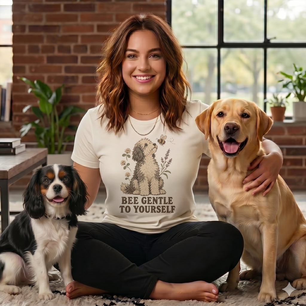 A smiling woman wearing a graphic t-shirt sits cross-legged, flanked by a Cavalier King Charles Spaniel and a Labrador Retriever.