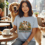 A woman sitting at a cafe table wearing a light blue t-shirt featuring a garden graphic and text that reads "GARDEN ASSISTANT, results may vary," with a golden retriever standing behind her.