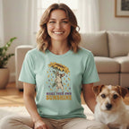 A smiling woman wears a mint green t-shirt with a French bulldog and sunflower graphic, sitting beside a terrier dog.