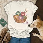Off-white crewneck t-shirt featuring a basket of colorful yarn balls with paw print designs, displayed with a dog, shoes, and jeans.