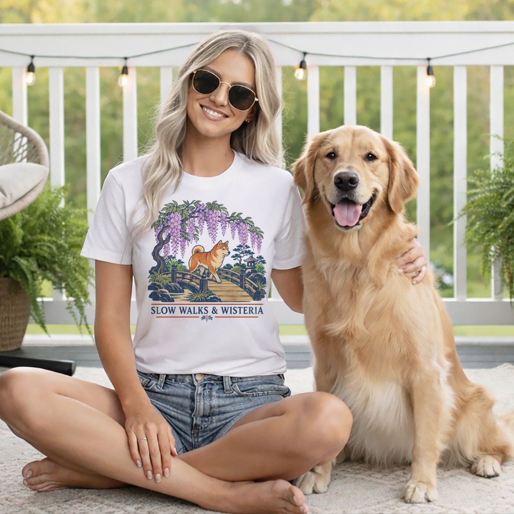 A smiling blonde woman in sunglasses sits beside a golden retriever, wearing a white t-shirt featuring a dog and wisteria illustration.