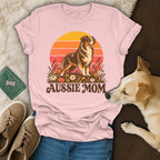A light pink t-shirt with an Australian Shepherd graphic and the text Aussie Mom rests on a wooden surface with a live dog.
