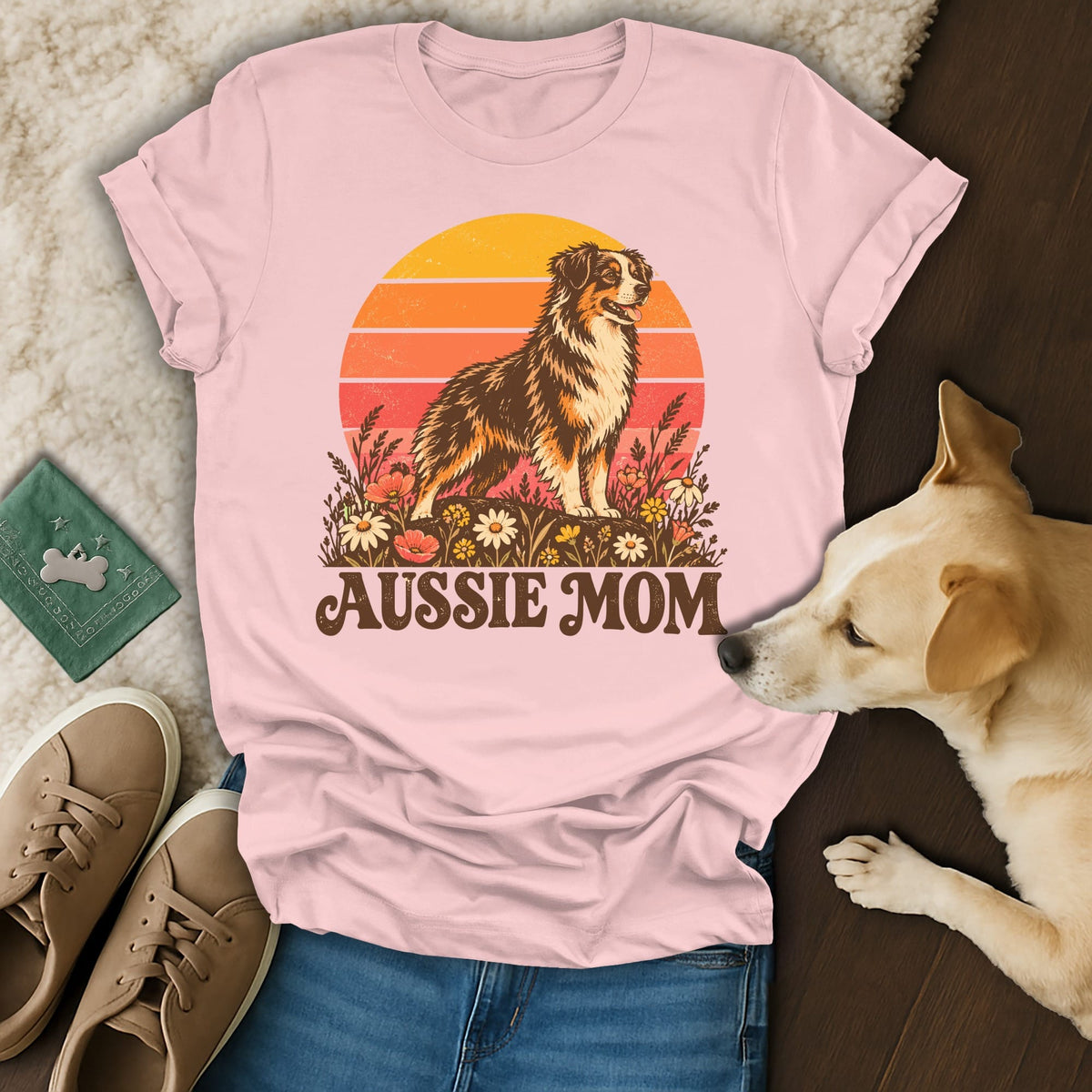 A light pink t-shirt with an Australian Shepherd graphic and the text Aussie Mom rests on a wooden surface with a live dog.