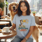A smiling woman wearing a light blue t-shirt with a dog graphic that says SPREAD SOME JOY sits next to a golden retriever at a cafe table with coffee.