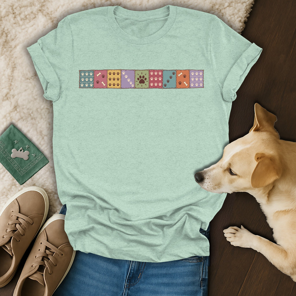 A light green t-shirt featuring a colorful paw print patchwork design across the chest, laid flat with jeans, sneakers, and a dog.