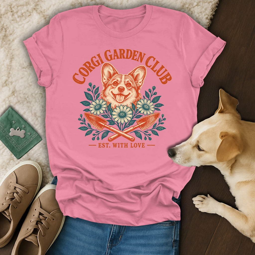 A pink t-shirt featuring a Corgi Garden Club graphic with a smiling corgi, flowers, and crossed garden trowels.