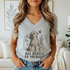 A person wearing a grey V-neck t-shirt featuring a graphic of a dog looking at a bee with the text BEE GENTLE TO YOURSELF.