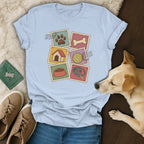 Light blue t-shirt featuring dog-themed postage stamps, laid flat with a dog, jeans, shoes, and accessories.