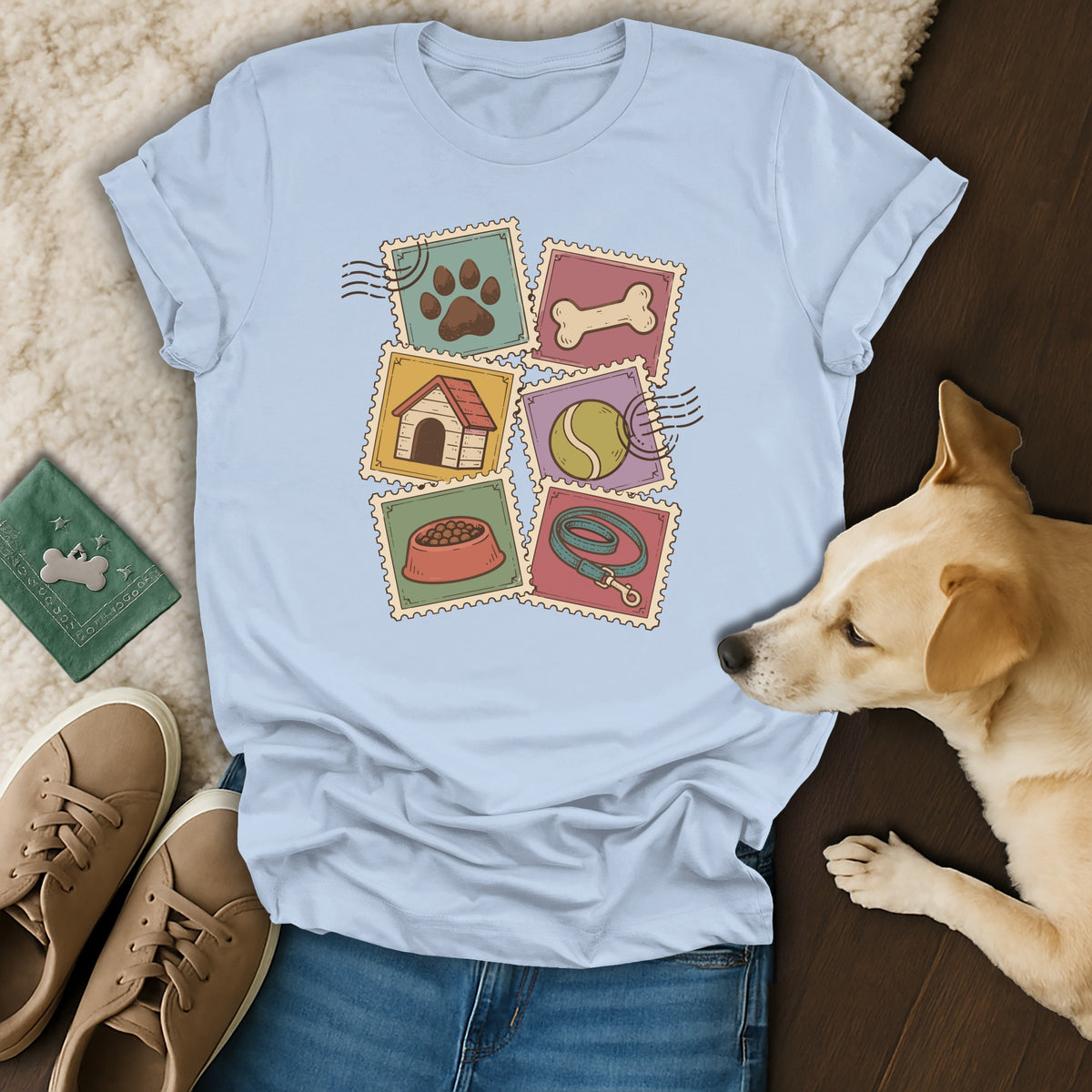 Light blue t-shirt featuring dog-themed postage stamps, laid flat with a dog, jeans, shoes, and accessories.
