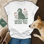 A white t-shirt with a green doodle dog graphic, floral elements, and the text GROWS BEST BESIDE YOU.