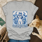 Gray V-neck t-shirt featuring a blue and white Delft-style illustration of a puppy, tulips, birds, and text reading Tulips & Tiny Paws.