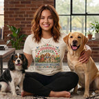 A smiling woman sitting cross-legged wearing a t-shirt with Grandmas Garden Granddogs Welcome printed on it, flanked by a Cavalier King Charles Spaniel and a Labrador Retriever.