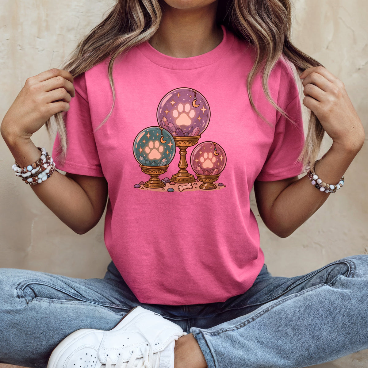 A woman wears a bright pink t-shirt printed with an illustration of three crystal balls featuring paw prints.