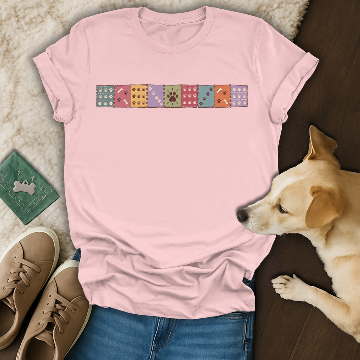 Pink t-shirt with a horizontal row of colorful patchwork squares featuring paw prints, alongside jeans, shoes, and a dog.