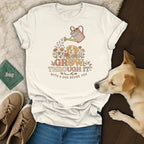 Cream t-shirt with graphic: dog, flowers, watering can, and text "GROW THROUGH IT WITH A DOG BESIDE YOU."