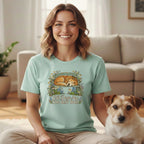 A smiling woman wears a mint green t-shirt with a vintage dog and flower graphic, sitting next to a small white and brown dog.