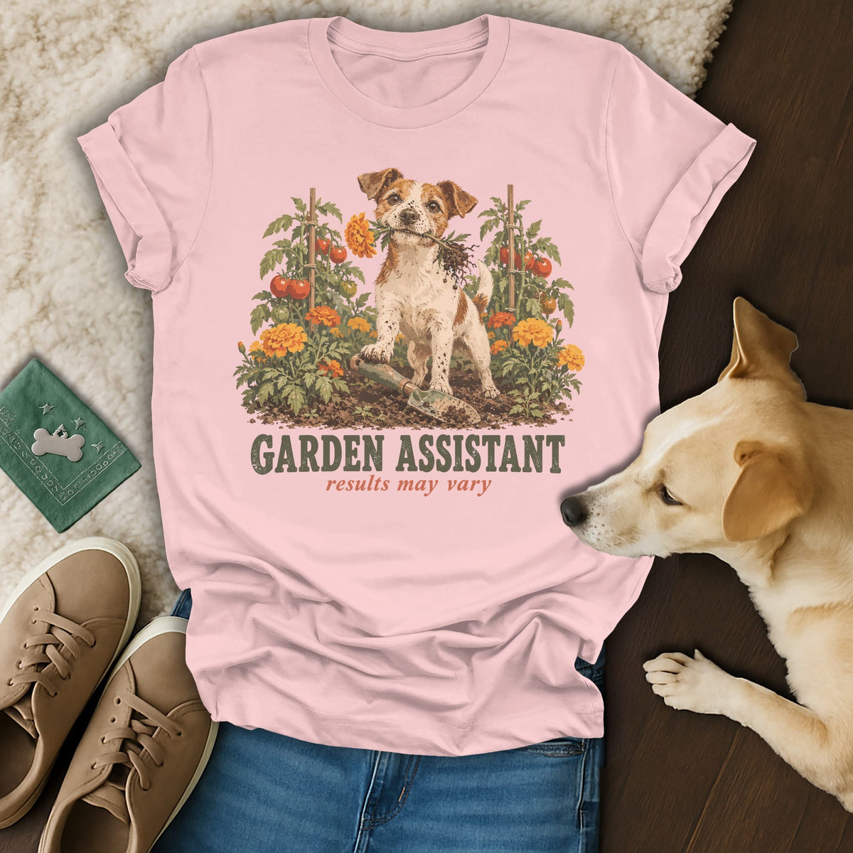 A light pink t-shirt with a graphic of a dog gardening, text reading GARDEN ASSISTANT results may vary, laid flat with jeans and shoes.
