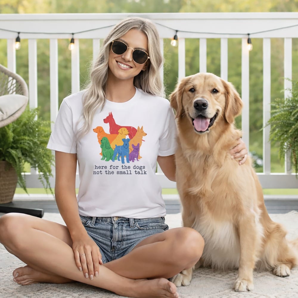 Woman in white t-shirt with rainbow dogs graphic and text saying "here for the dogs not the small talk" sits with golden retriever.