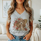 A woman wears a grey V-neck t-shirt featuring a graphic of a cockapoo dog in a flower-adorned hat, with the text Garden Party guest of honor below.