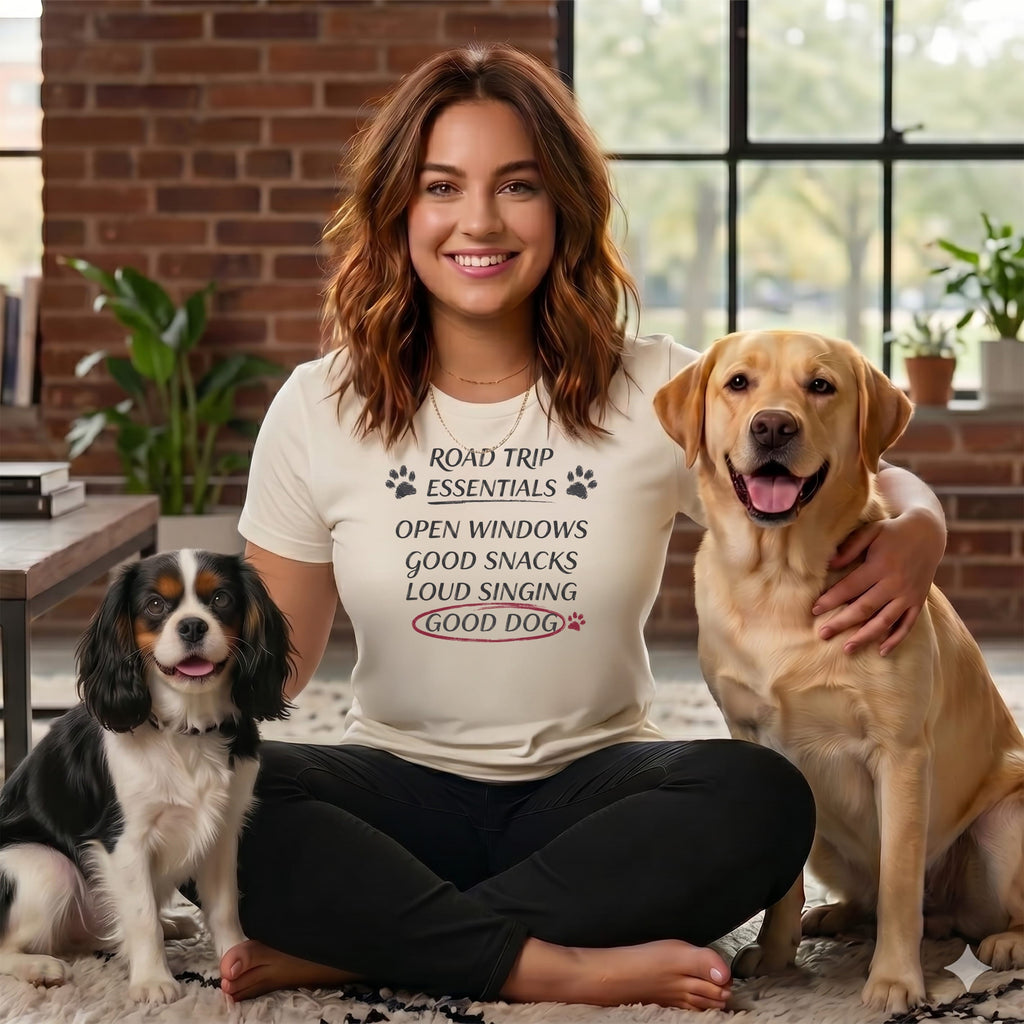 Woman with two dogs wearing t-shirt that says "Road Trip Essentials: Open Windows, Good Snacks, Loud Singing, Good Dog".