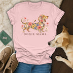 A pink t-shirt with a floral dachshund graphic and the text DOXIE MAMA lies on a white blanket next to blue jeans, brown shoes, and a real dog.