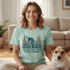A smiling woman wears a mint green t-shirt with a sniffing dog illustration and the text FOLLOW YOUR NOSE TO THE FLOWERS, sitting next to a small terrier.