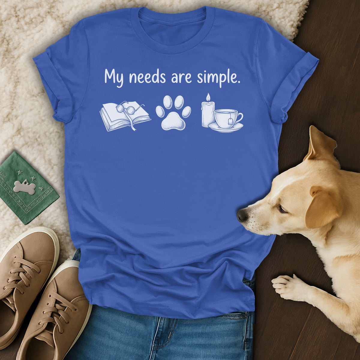 Blue t-shirt with "My needs are simple" text and icons of book, paw, candle, and tea.