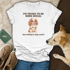 White t-shirt with a drawing of a dog holding a cup, text says "I'M TRYING TO BE MORE SOCIAL does talking to dogs count?"