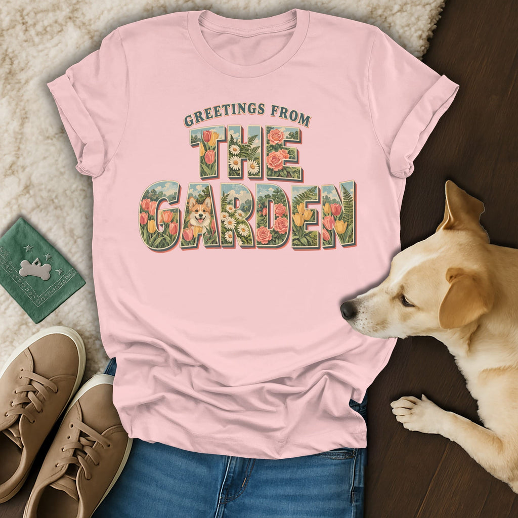Pink crewneck t-shirt featuring a vintage floral graphic with the text Greetings From The Garden, laid on a fuzzy blanket next to a dog, shoes, and jeans.