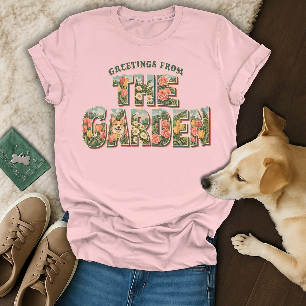 Pink crewneck t-shirt featuring a vintage floral graphic with the text Greetings From The Garden, laid on a fuzzy blanket next to a dog, shoes, and jeans.
