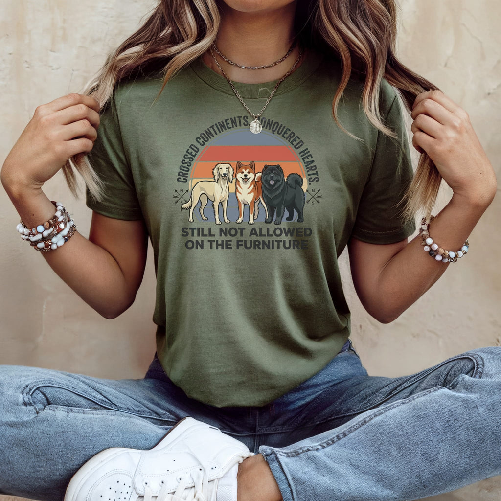 Green t-shirt with three dogs and sunset graphic; text reads "Crossed Continents, Conquered Hearts, Still Not Allowed on the Furniture."