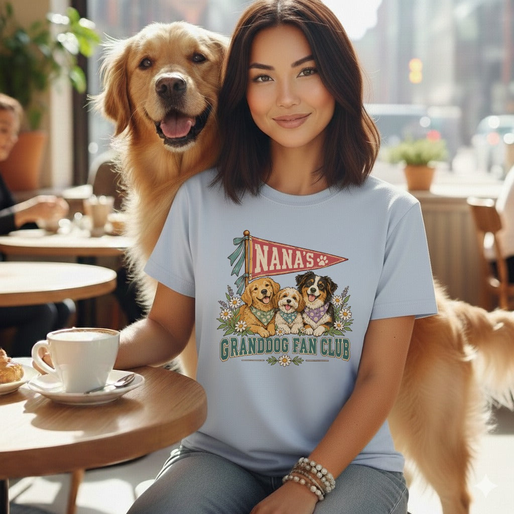 A woman wearing a light blue t-shirt with a graphic of dogs and text that reads Nana's Granddog Fan Club sits at a table with a golden retriever standing behind her.