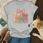 Light gray crewneck t-shirt featuring a pastel stack of bows with paw prints, surrounded by casual items and a small dog.